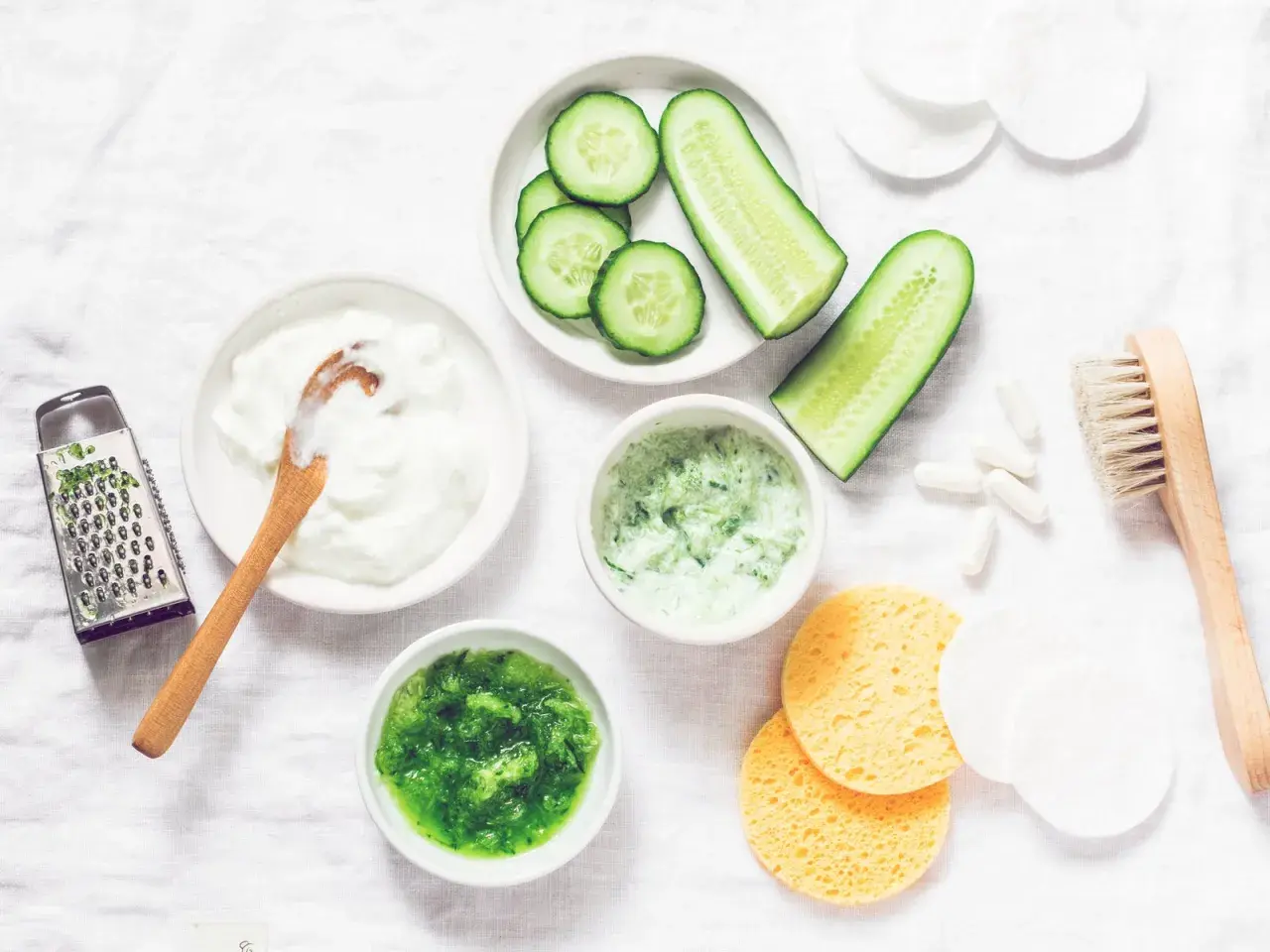 Zutaten für eine Gurkenmaske selber machen: Gurkenscheiben, Joghurt, geriebene Gurke, ein Hobel und weitere Utensilien.