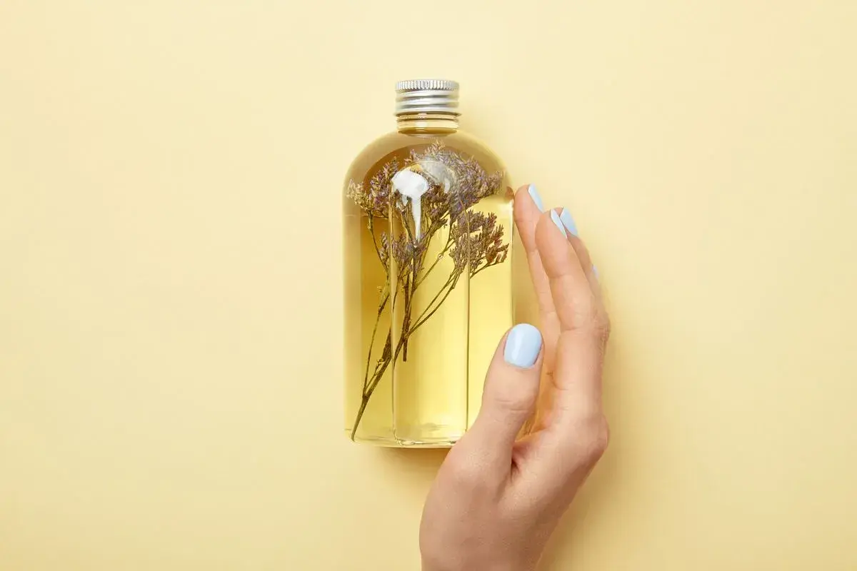 Eine Hand mit hellblauen Nägeln berührt eine Flasche mit gelbem Öl und getrockneten Blumen. Perfekt für duschgel selber machen ohne seife.