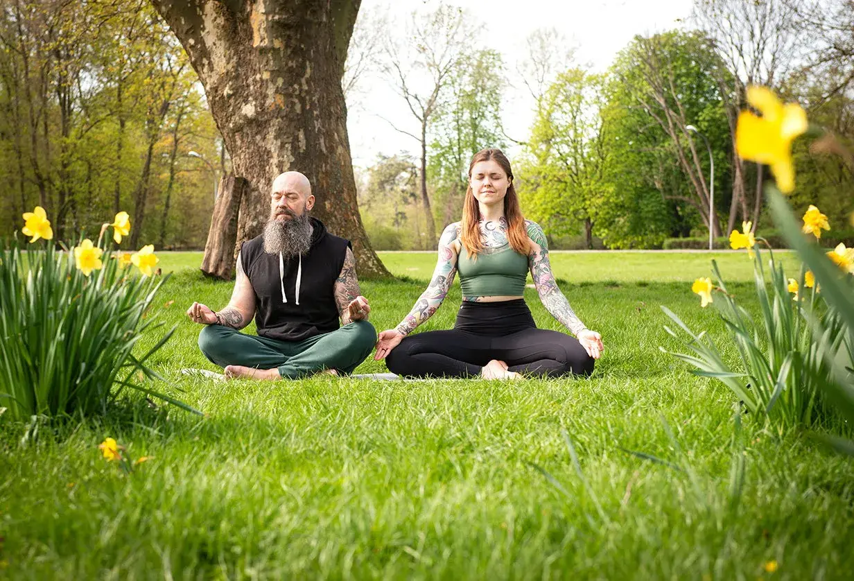 Mann und Frau in bequemer Kleidung machen Meditation im Sitzen auf einer Wiese, umgeben von Narzissen und Bäumen.
