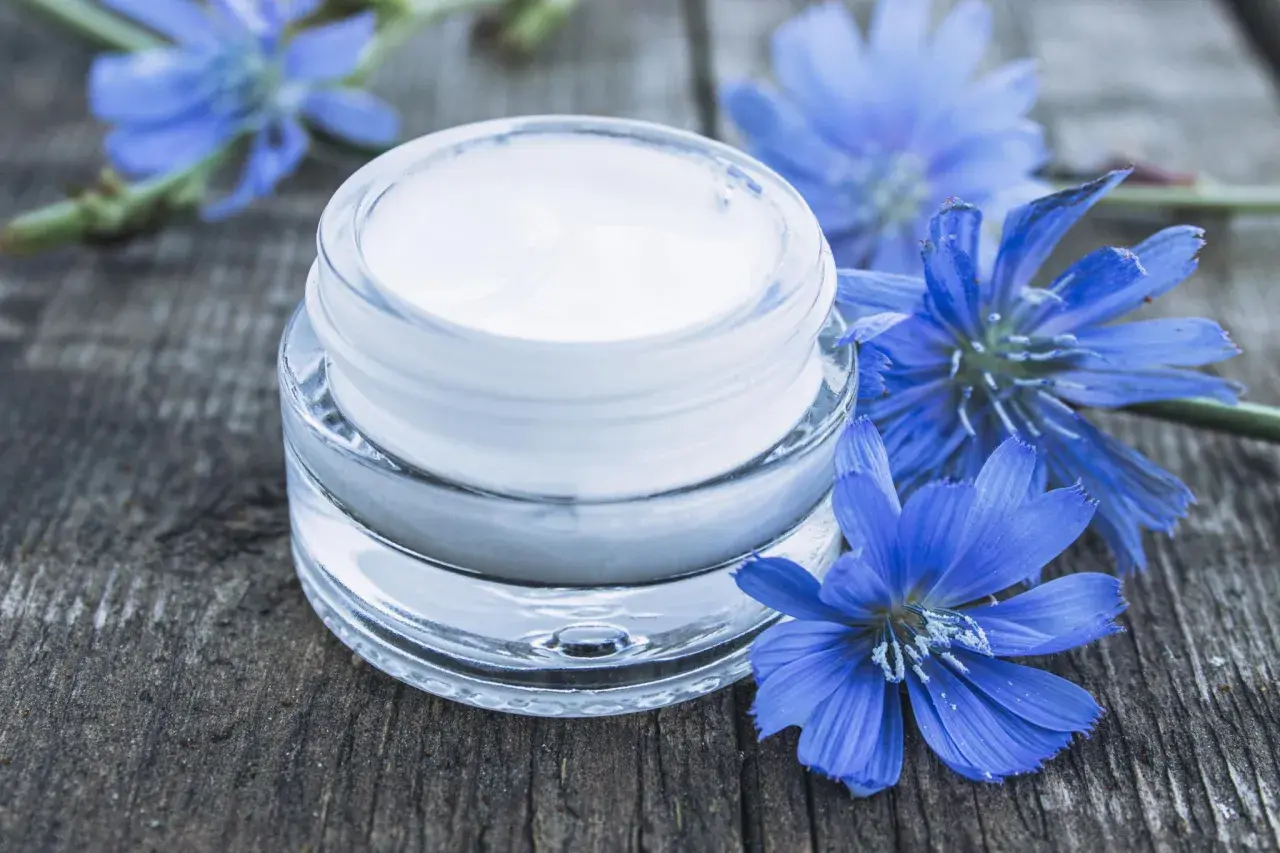 Creme für trockene Haut: Ein Glas mit weißer Creme neben blauen Wegwartenblüten auf Holz. Perfekt zum gesichtscreme selber machen trockene haut.