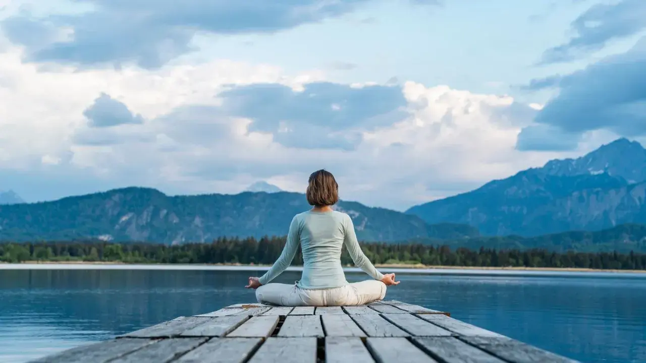 Frau in Lotusposition auf einem Steg, genießt eine einfache Meditation mit Blick auf einen Bergsee und die Alpen.