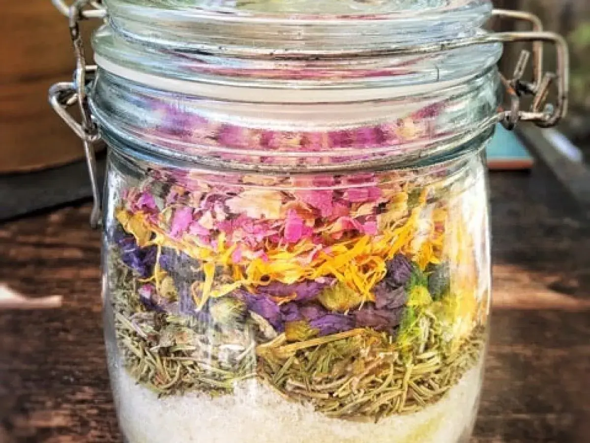 Ein Glas mit selbstgemachtem Badezusatz: unten Salz, darauf Rosmarin, dann lila Blüten und oben rosa Blütenblätter.