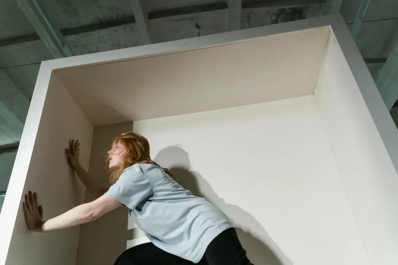 Eine Frau mit roten Haaren stemmt sich gegen eine Wand, als wolle sie die Opferrolle verlassen und aus einer Begrenzung ausbrechen.