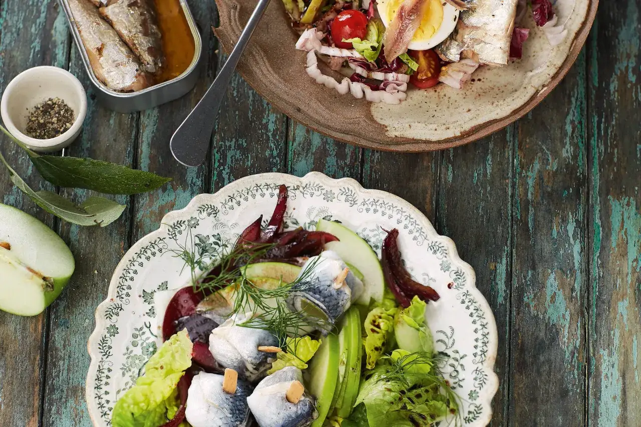 Zwei Teller mit sardinen salat, einer mit Roter Bete und Apfel, der andere mit Ei und Tomaten.