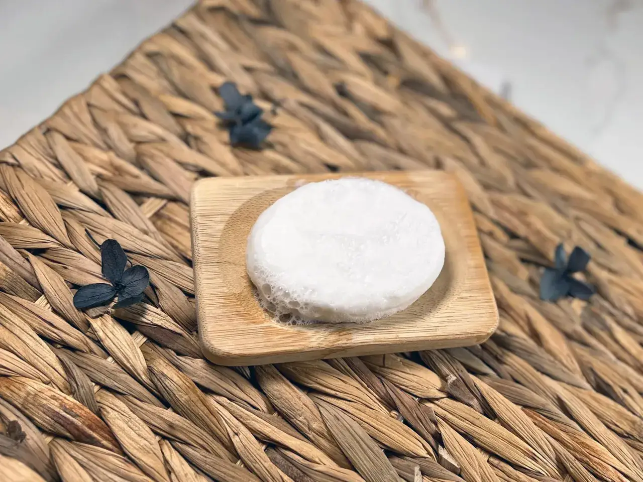 Ein selbstgemachtes Duschbar-Rezept ist einfach! Hier ist ein Beispiel: ein wei&szlig;er, runder Seifenblock auf einem Holzbrett, umgeben von getrockneten Blumen auf einem geflochtenen Untergrund.