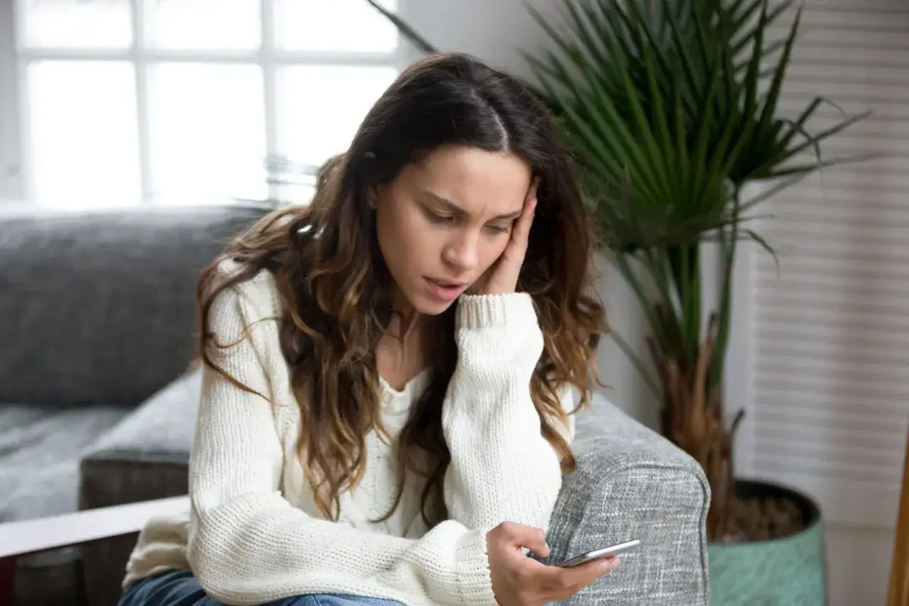 Eine Frau sitzt nachdenklich auf einem Sofa und hält ihr Handy. Sie fragt sich, wie werde ich negative gedanken los?