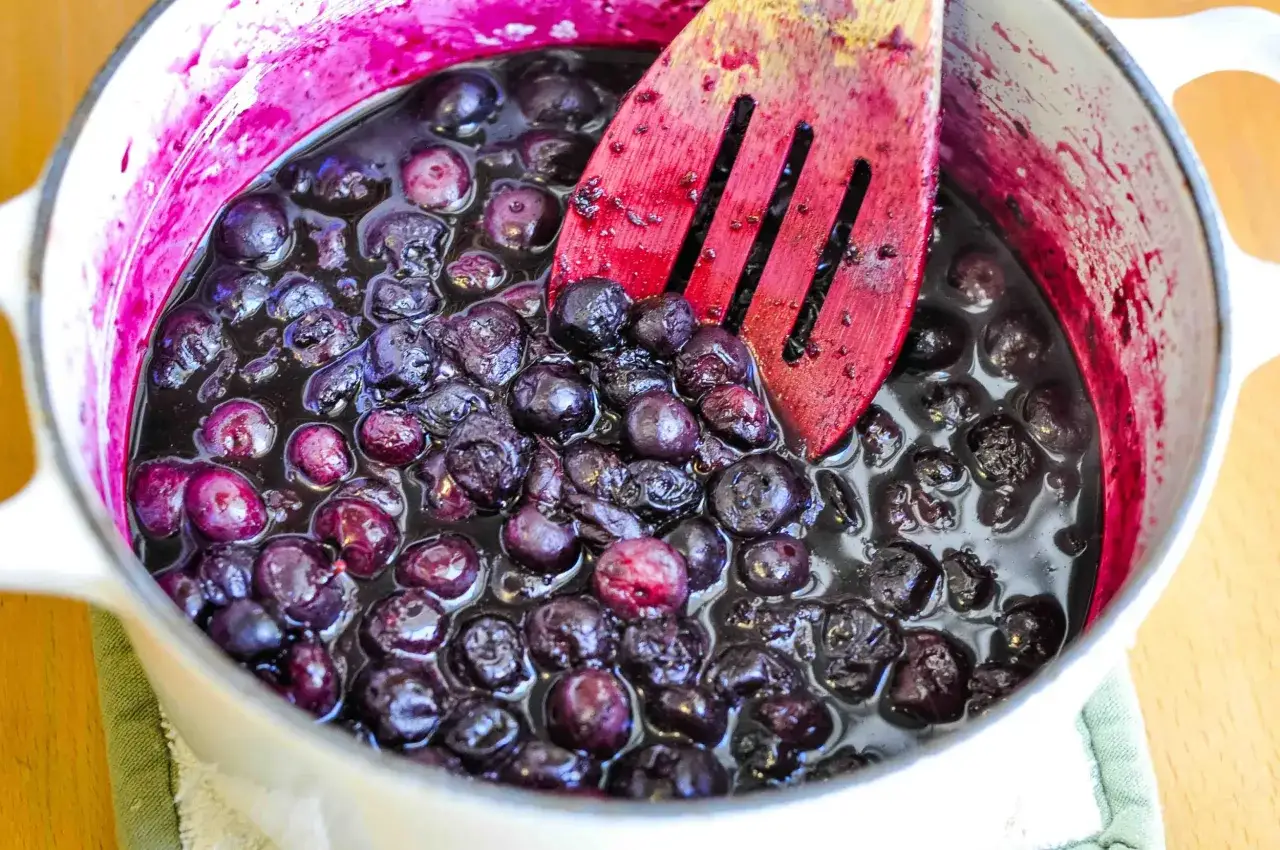 Heidelbeeren kochen mit Zucker in einem Topf, eine rote Holzkelle r&uuml;hrt die s&uuml;&szlig;e Masse.