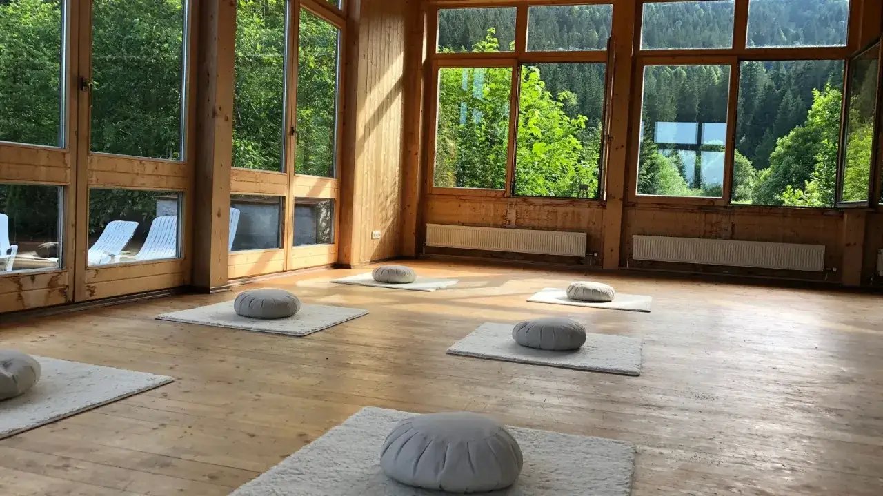 Ein heller Raum mit Holzboden und gro&szlig;en Fenstern, die auf einen gr&uuml;nen Wald blicken. Mehrere Meditationskissen laden zum schweigen statt reden ein.