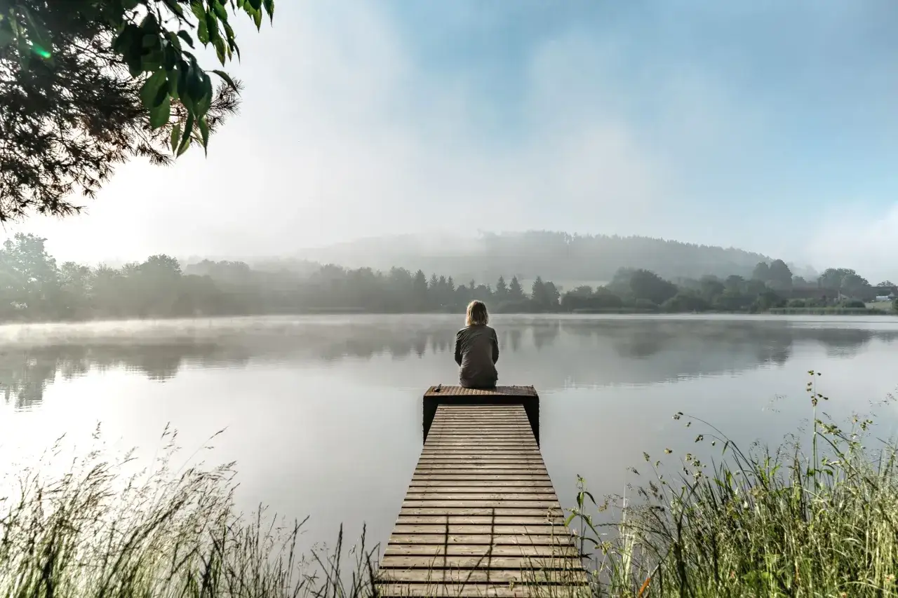 Eine Person sitzt auf einem Steg am See und genießt die Ruhe. Die Nebelschwaden über dem Wasser laden dazu ein, weniger Gedanken zu machen.