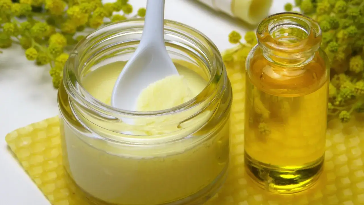 Glas mit Honigcreme und Löffel, daneben kleines Glas mit goldenem Öl, gelbe Blüten im Hintergrund.