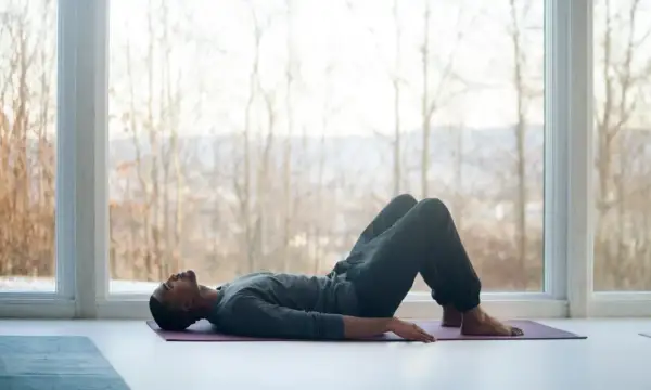 Mann liegt auf Yogamatte, Knie angewinkelt, entspannt. Fokus auf tiefe meditation atmung vor großem Fenster mit Blick auf kahle Bäume.