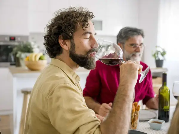 Mann genießt ein Glas Rotwein am Abend, während ein Freund im Hintergrund sitzt.
