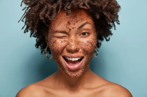 Frau mit braunem Peeling im Gesicht, die zwinkert und lacht. Ein tolles Beispiel, wie man ein Heilerde Peeling selber machen kann.