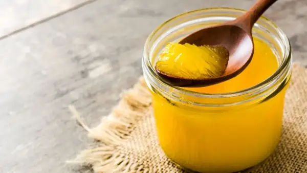 Ein Glas mit goldgelbem Ghee, das auf einem Holzlöffel entnommen wird. Ghee ist geklärte Butter, ein vielseitiges Fett.