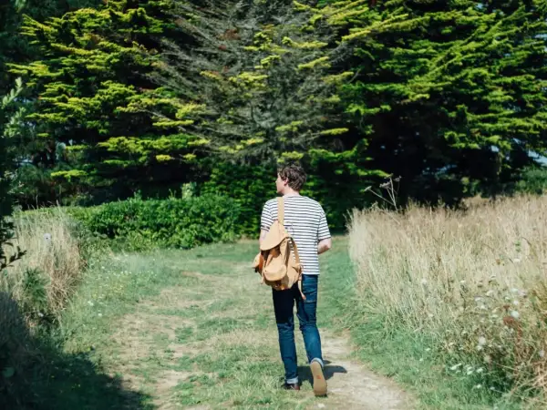 Mann mit Rucksack auf einem Feldweg, umgeben von grünen Bäumen und Gräsern. Ein Moment des meditativen Gehens in der Natur.
