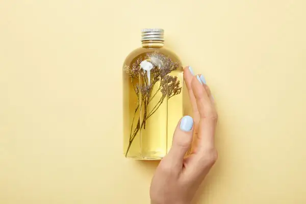 Eine Hand mit hellblauen Nägeln berührt eine Flasche mit gelbem Öl und getrockneten Blumen. Perfekt für duschgel selber machen ohne seife.