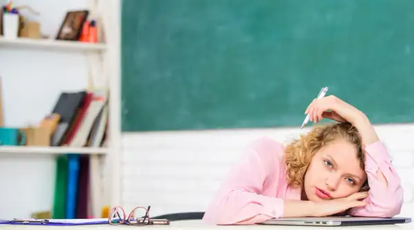 Junge Lehrerin mit Locken, die mit einem Stift auf dem Schoß liegt, wirkt gestresst. Hinter ihr eine Tafel und ein Regal voller Bücher.